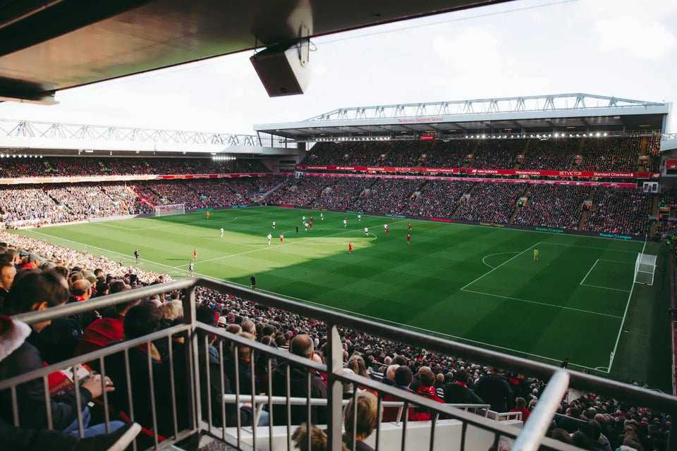 Stade de football rouge et blanc avec une grande zone de sièges, des balustrades en métal, un terrain herbeux et un tableau de scores.